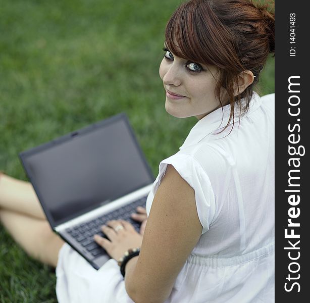 Pretty young woman working on a laptop computer outdoors, lying on green grass, pondering, looking away