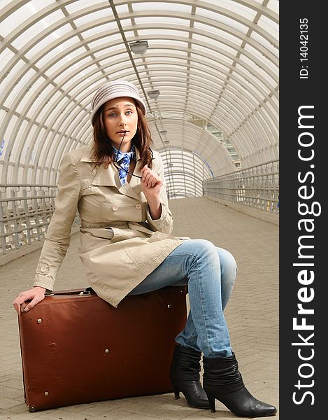 The young beautiful girl at station with a suitcase in a hand, in a light hat and a raincoat