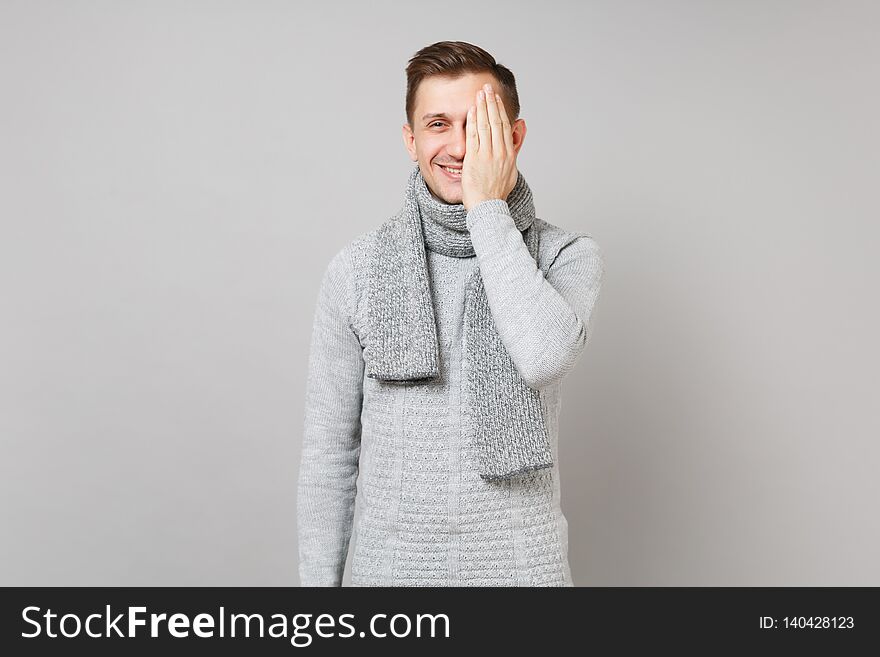 Smiling young man in gray sweater, scarf covering eyes with hand on grey wall background in studio. Healthy fashion lifestyle people sincere emotions, cold season concept. Mock up copy space