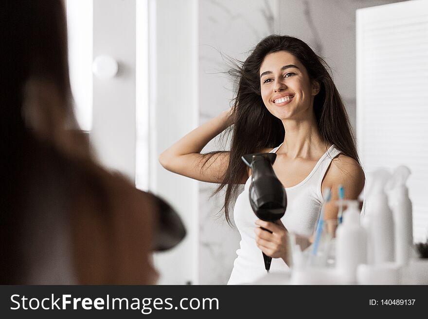Smiling beautiful girl drying her hair in front of mirror in bathroom.