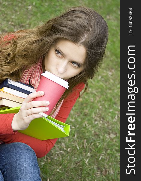 Young beautiful student at a dinner in the park sits on the green grass drinks coffee. Keep lots of books and folders. Young beautiful student at a dinner in the park sits on the green grass drinks coffee. Keep lots of books and folders