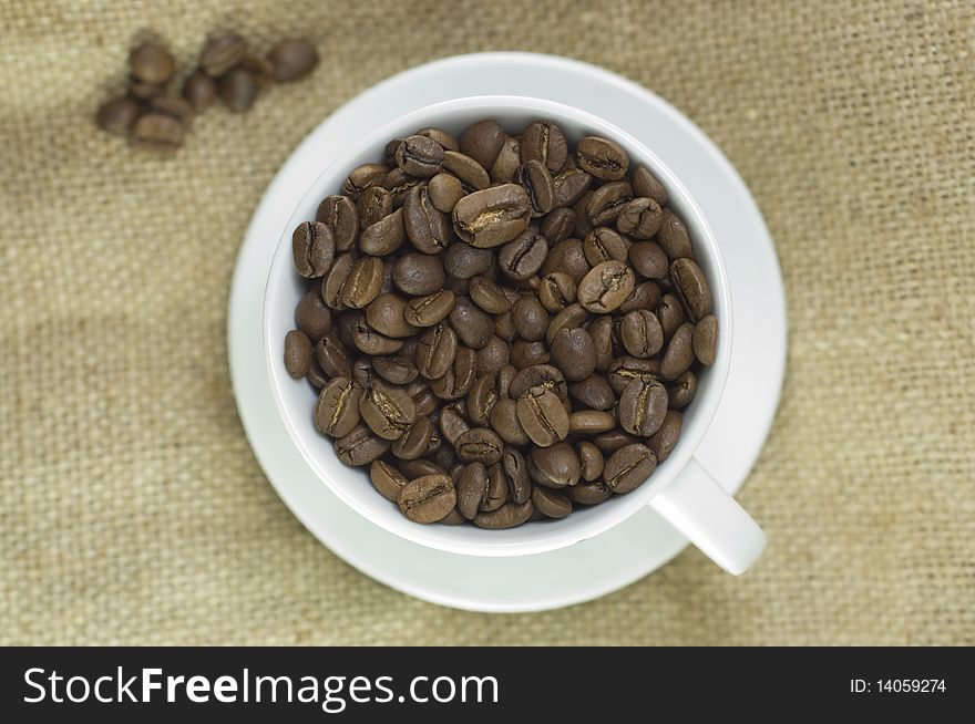 Roasted coffee beans in white cup, on a linen bag, macro. Roasted coffee beans in white cup, on a linen bag, macro