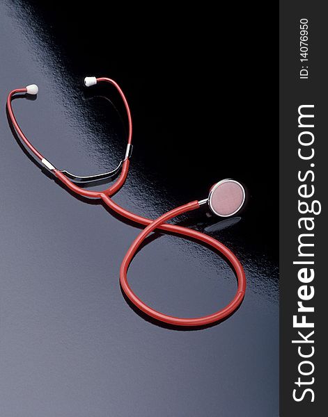 A red stethoscope is laying on a black reflective surface. Vertical shot.