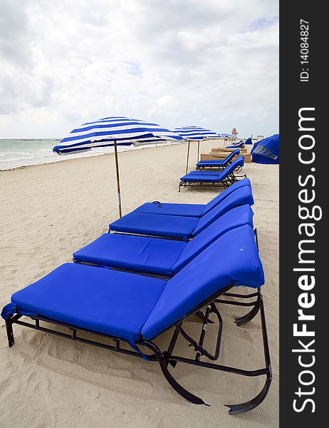 Row of blue beach lounge chairs and matching umbrellas along the shoreline of Miami Beach. Row of blue beach lounge chairs and matching umbrellas along the shoreline of Miami Beach.