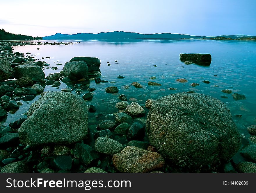 Boulders, cobble, rocks and stones on a beach are exposed at low tide in a calm inlet with distant shore and morning sky background. Boulders, cobble, rocks and stones on a beach are exposed at low tide in a calm inlet with distant shore and morning sky background.