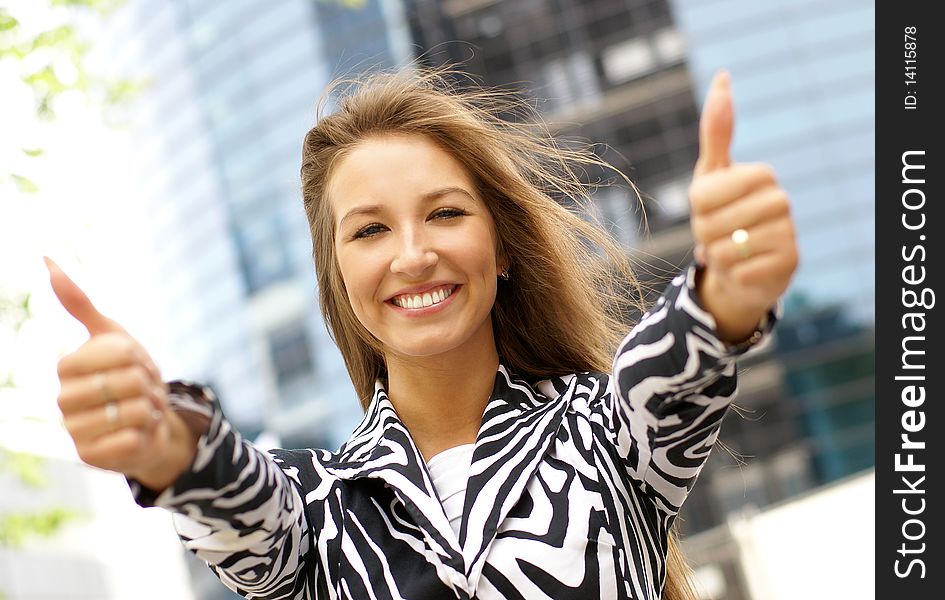 Portrait on a young business woman holding thumbs up as a sign of success. Image taken on a outdoors, on a modern background.