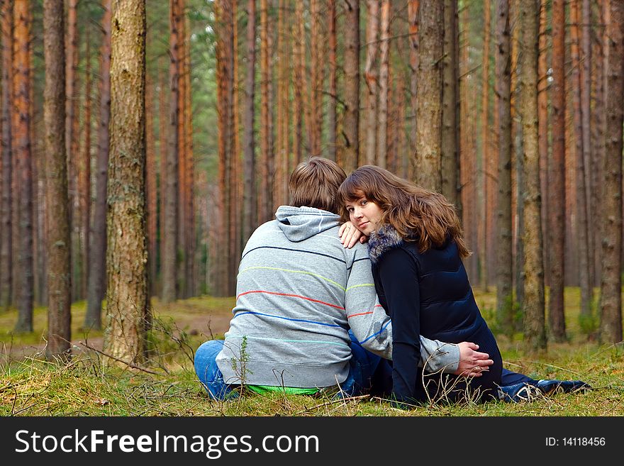 Young loving couple sitting back on a grass in the forest, woman is turning her face, they are hugging each other and dreaming. Young loving couple sitting back on a grass in the forest, woman is turning her face, they are hugging each other and dreaming