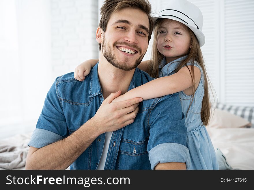 Portrait of handsome father and his cute daughter hugging, looking at camera and smiling while sitting on sofa at home.