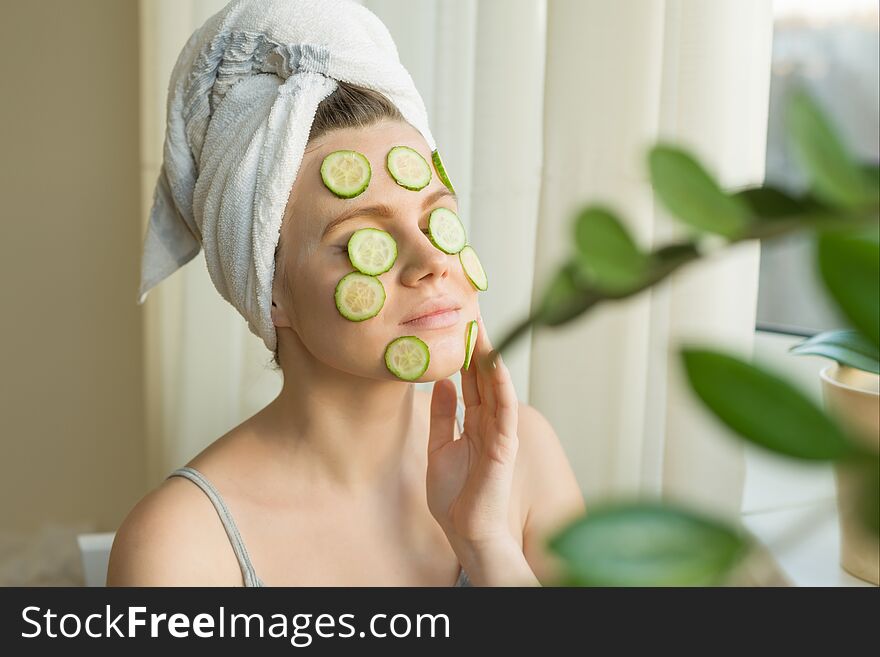 Young beautiful woman close-up at home near the window with natural homemade mask of cucumber on face, towel on head. Skin care