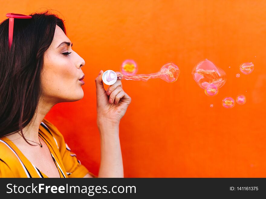 Side view shot of young beautiful female blowing soap bubbles against orange background. Young woman having fun. Side view shot of young beautiful female blowing soap bubbles against orange background. Young woman having fun