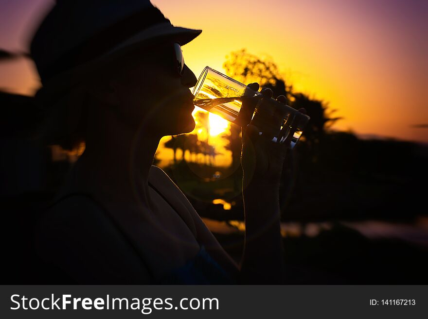 Woman with white hat and pink sunglasses with nice reflection of palm trees and sunset drinking fresh pure water