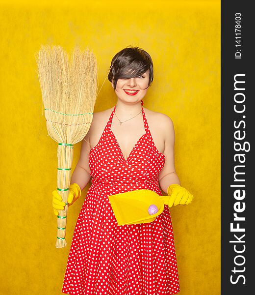 Cute charming emotional young woman in red retro polka dot dress stands in rubber gloves for cleaning with a broom in her hands fo