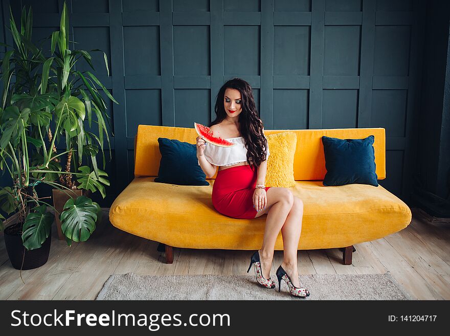 Stylish pregnant woman sitting on orange sofa, eating watermelon and posing. Attractive future mother in elegant clothes awaiting for little baby and relaxing. Concept of pregnancy and fashion. Stylish pregnant woman sitting on orange sofa, eating watermelon and posing. Attractive future mother in elegant clothes awaiting for little baby and relaxing. Concept of pregnancy and fashion.