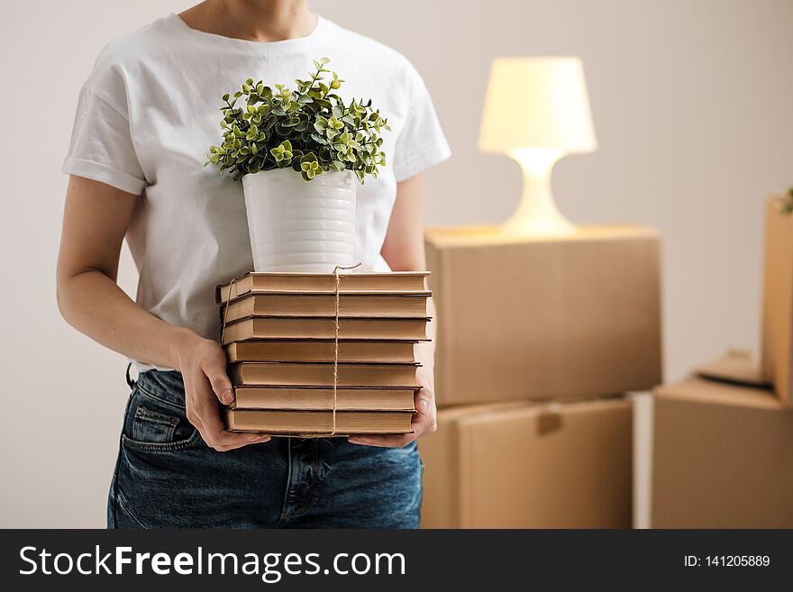 The concept of relocation and moving to a new home. Close-up, female hands hold a pile of books and a green plant in a pot. On the background of cardboard boxes and a table lamp.