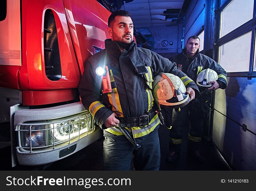 Two firemen wearing protective uniform standing next to a fire truck in a garage of a fire department.