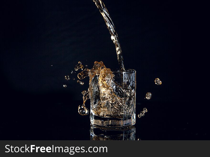 Whiskey pouring into glass with ice isolated on reflective black surface. Whiskey splash out of glass, many drops of beverage get out from glass. Whiskey pouring into glass with ice isolated on reflective black surface. Whiskey splash out of glass, many drops of beverage get out from glass