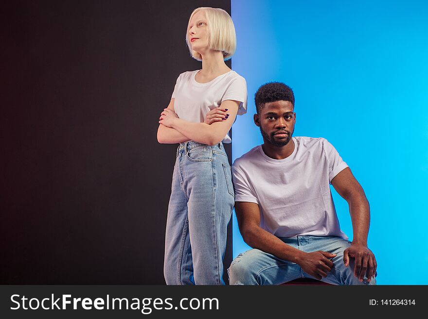 Attractive Afro men is sitting on the chair and looking at the camera, while his Caucasian albino girlfriend standing with crossed arms bear him, copy space. Attractive Afro men is sitting on the chair and looking at the camera, while his Caucasian albino girlfriend standing with crossed arms bear him, copy space
