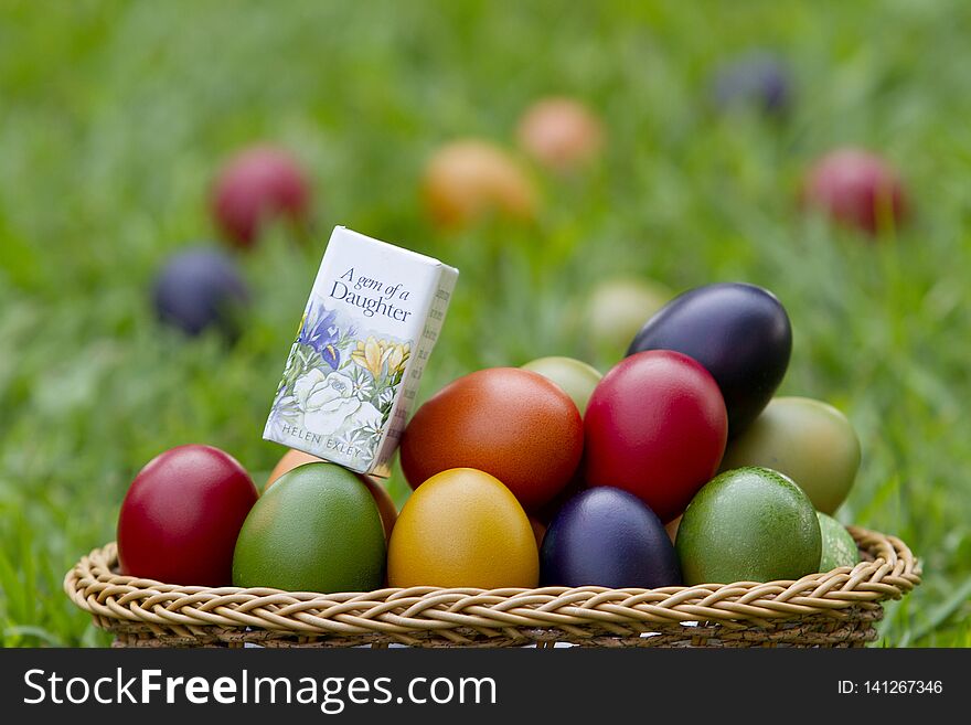 A basket of red, blue, green, orange and yellow painted eggs with a mini book, A gem of a daughter in some grass on a summer afternoon. A basket of red, blue, green, orange and yellow painted eggs with a mini book, A gem of a daughter in some grass on a summer afternoon