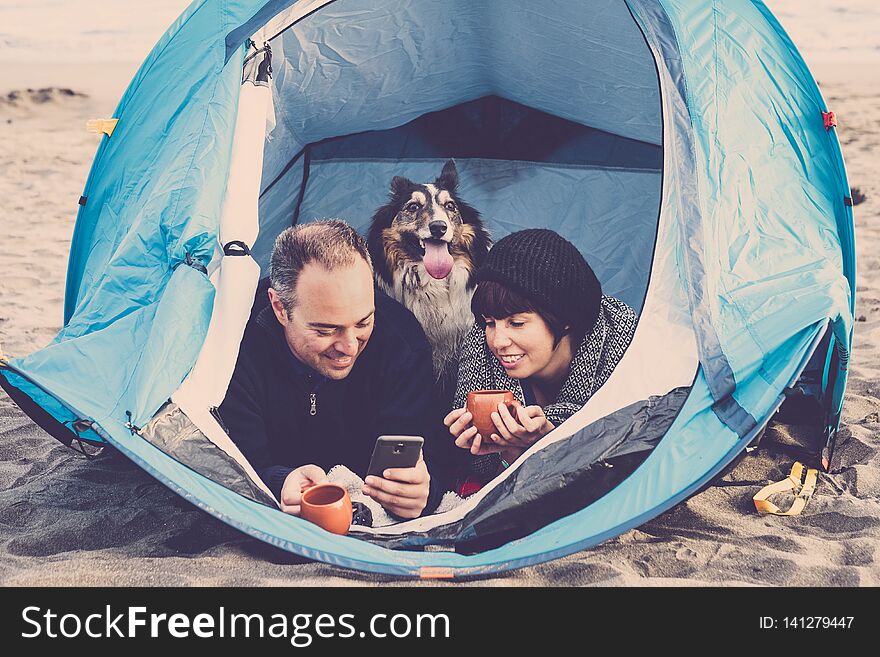 Couple looking at the smart phone and have fun inside a tent in free camping on the beach Dog border collie behind them looking at the camera. vintage colors and vacation family concept. alternative travel lifestyle. Couple looking at the smart phone and have fun inside a tent in free camping on the beach Dog border collie behind them looking at the camera. vintage colors and vacation family concept. alternative travel lifestyle