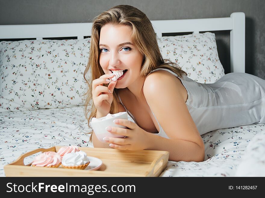 Young blonde with cupcakes eating marshmallow lying in bed