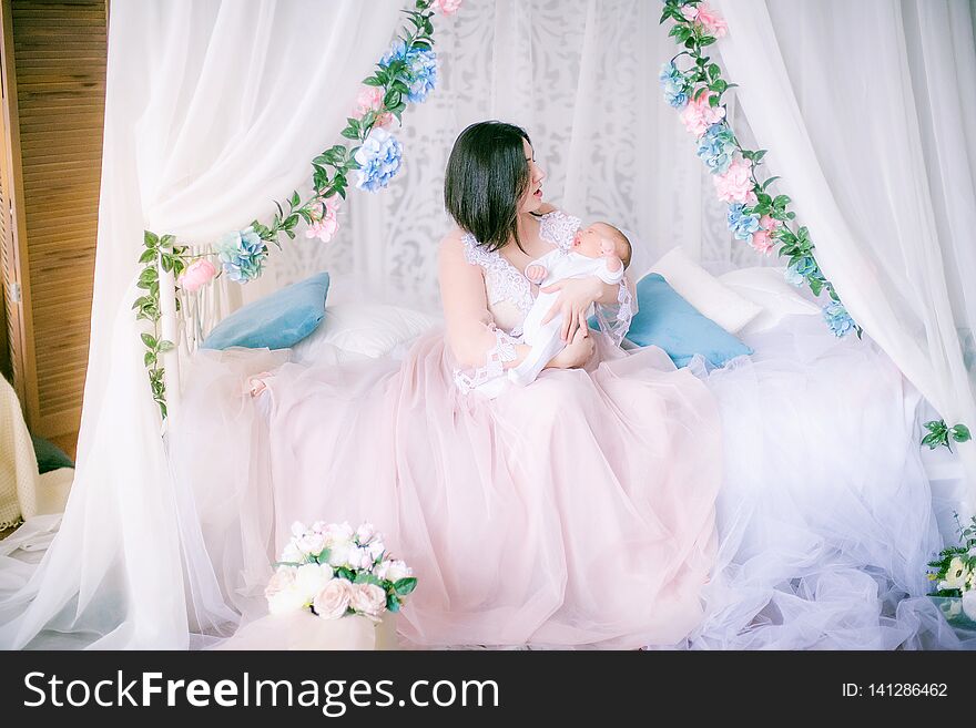 Young mother in a boudoir dress with a baby in her arms in spring flowers by the canopy bed. Young mother in a boudoir dress with a baby in her arms in spring flowers by the canopy bed