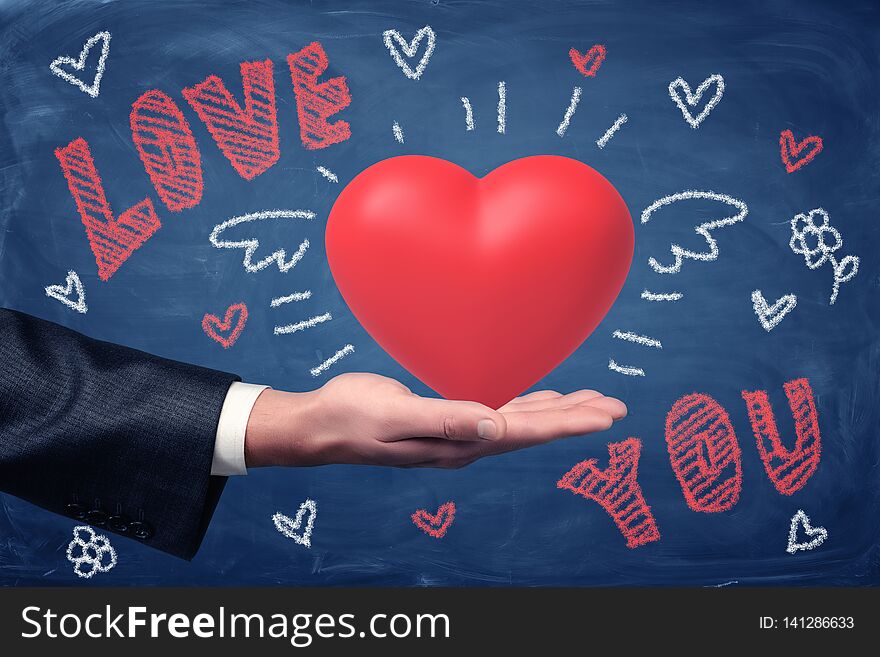 Male hand holding red heart with LOVE YOU chalk sign and heart drawings on blue background