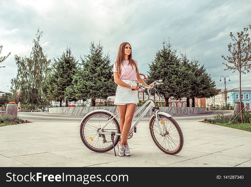 A Girl In A White Skirt And A Pink Blouse, Stands In Summer In The City Outdoors On A Weekend, A Parked Bicycle, Poses