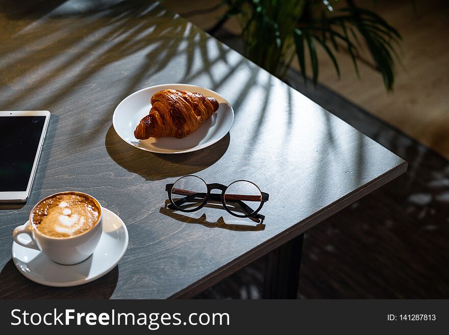 Close up of white coffee cup, croissant, glasses and pad with blank screen on dark wooden table with tropic plant on background. Close up of white coffee cup, croissant, glasses and pad with blank screen on dark wooden table with tropic plant on background