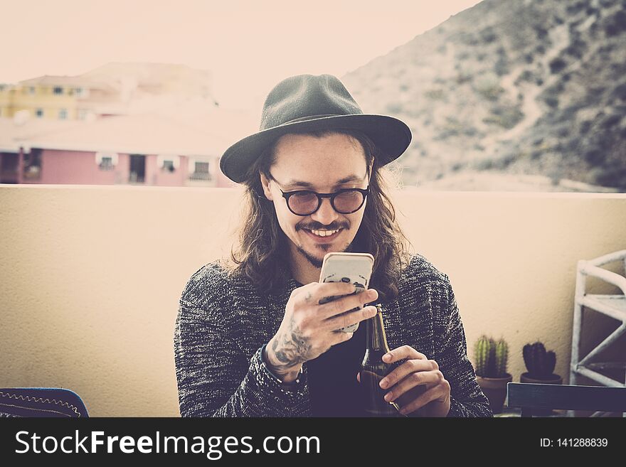 Long hair nice young man read and check the phone on the roofteop terrace outdoor. black hat and eyeglasses and tattoo on hand. fashion model sith nice smile. modern hipster concept. Long hair nice young man read and check the phone on the roofteop terrace outdoor. black hat and eyeglasses and tattoo on hand. fashion model sith nice smile. modern hipster concept