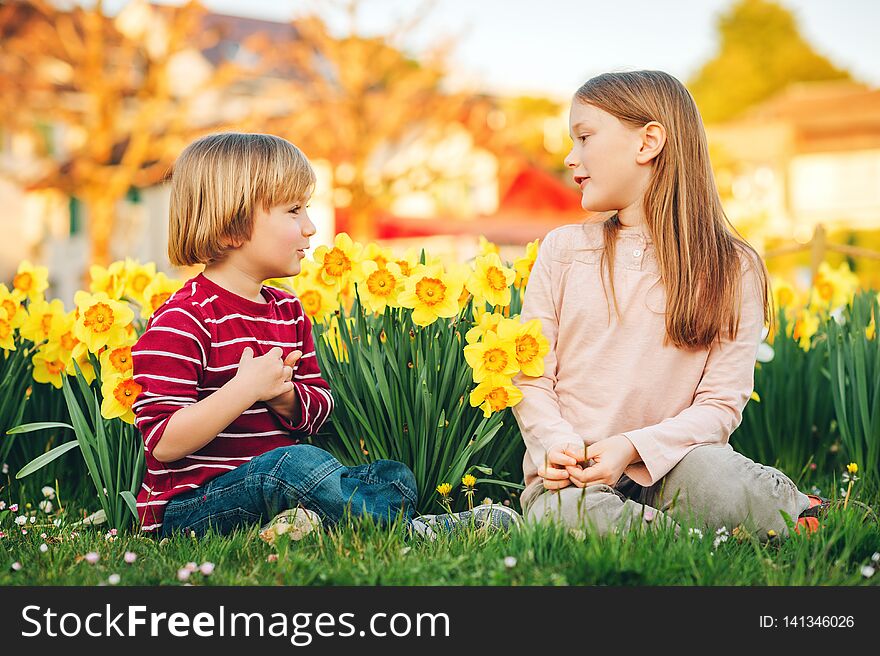 Two cute kids, little boy and his big sister, playing in the park between yellow daffodils flowers in sunlight