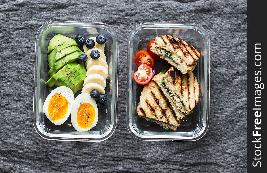 Two healthy office lunch box with sweet and savoury food. Boiled egg, avocado, tuna spinach cheese sandwiches, fruit on grey