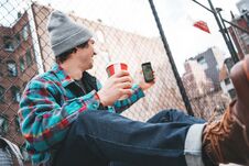 Young Trendy Man Sitting On The Street Sidewalk Resting With Cup Of Hot Coffee And Using Smartphone For Video Link Royalty Free Stock Photos