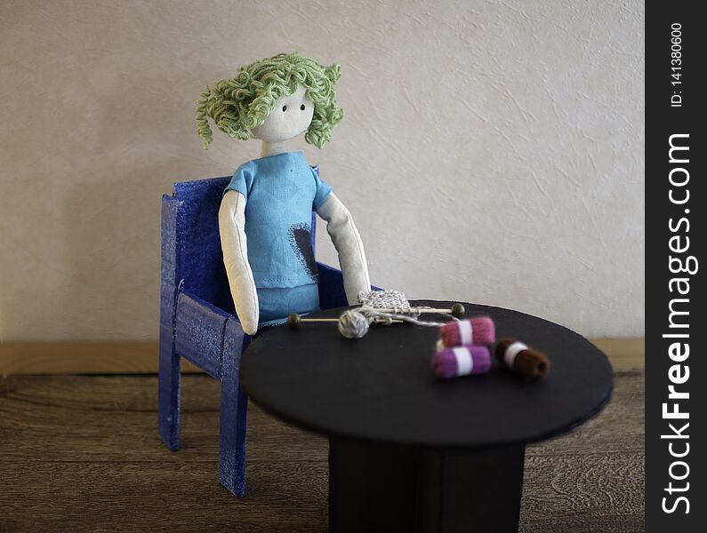Knitting image. A handmade rag doll is sitting at a black table with a knitting project with wooden knitting needles. Skeins of wool on the table. Room for copy. Knitting image. A handmade rag doll is sitting at a black table with a knitting project with wooden knitting needles. Skeins of wool on the table. Room for copy.