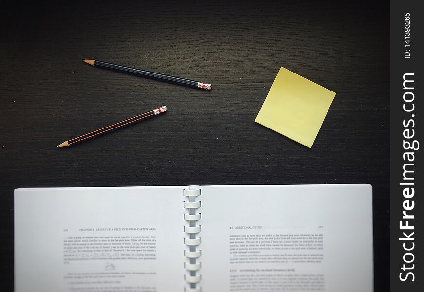 Textbook &#x28;blurred&#x29;, pencils, and yellow sticky note, on the dark wooden table. Textbook &#x28;blurred&#x29;, pencils, and yellow sticky note, on the dark wooden table