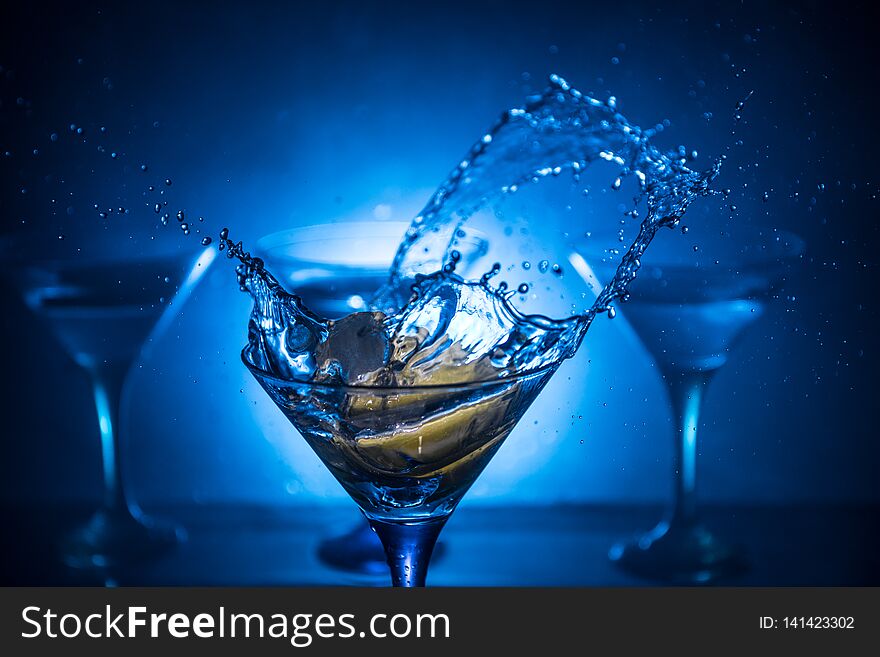 Martini cocktail glass in hand splashing on dark toned smoky background or colorful cocktail in glass with splashes and olives.
