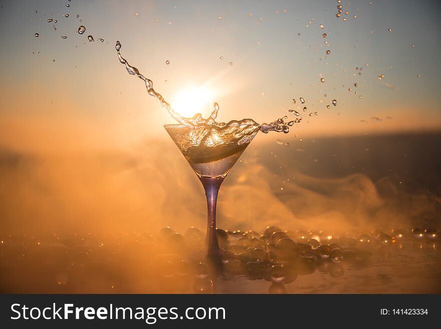 Martini cocktail glass splashing on foggy sunset background or colorful cocktail in glass with splashes and lemon. Outdoor open air party entertainment. Selective focus. Martini cocktail glass splashing on foggy sunset background or colorful cocktail in glass with splashes and lemon. Outdoor open air party entertainment. Selective focus