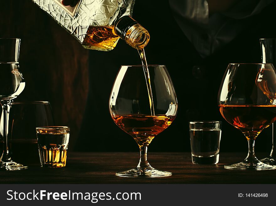 The Bartender Pours The Cognac Or Brandy In Big Wine Glass On The Old Bar Counter. Vintage Wooden Background In Pub Or Bar, Night