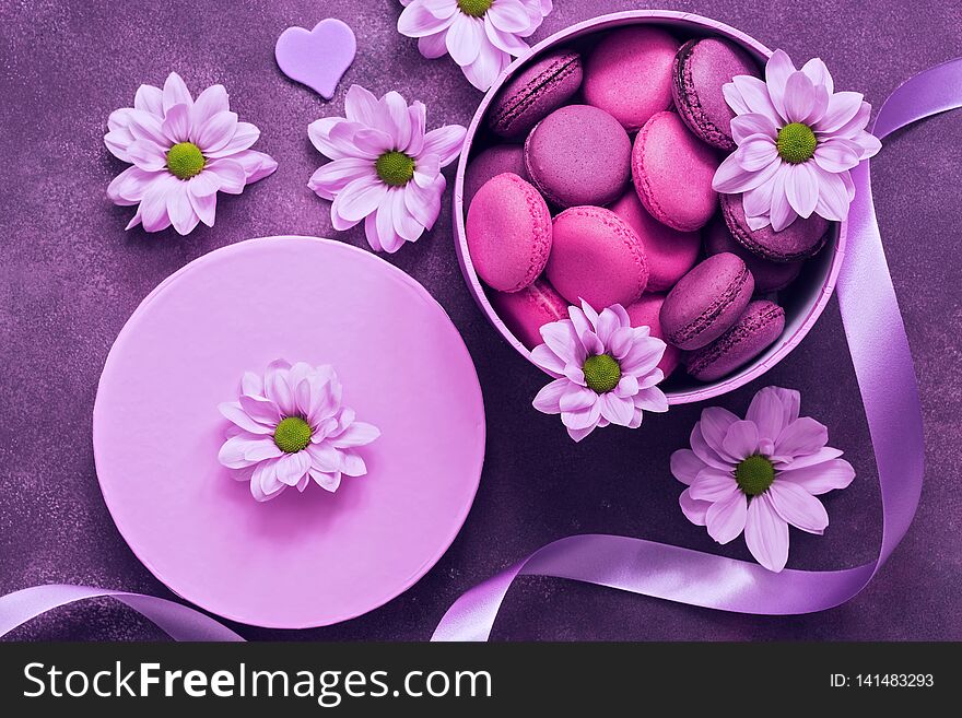 Purple and pink macaroons in a gift box on a beautiful purple background decorated with flowers. Top view