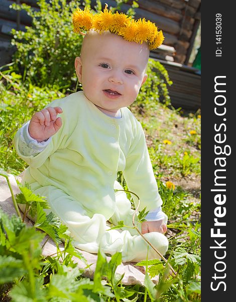 The baby of 7-8 months sits on a grass with a wreath from dandelions on a head and waves discontentedly hands