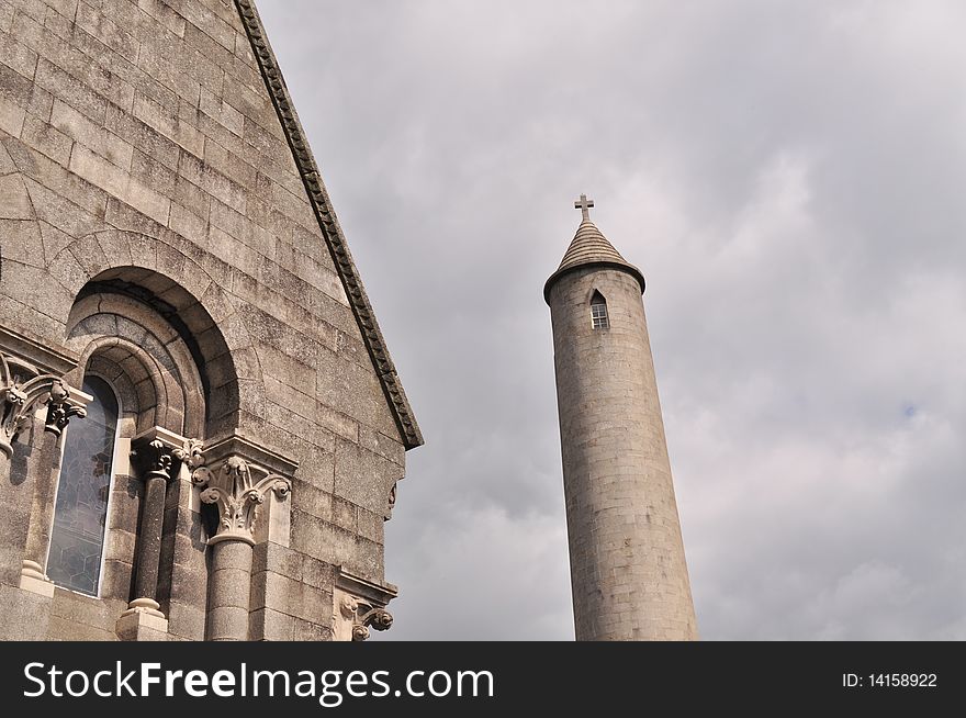 Image of a tall graveyard tower with window on top and section of small church with arch window in cemetery in north dublin ireland on cloudy day.