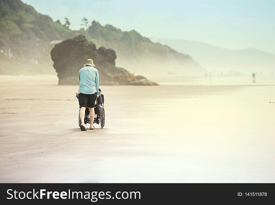 Father pushing disabled young child in wheelchair along sandy beach by the ocean on foggy misty day, walking away from the camera, back view. Father pushing disabled young child in wheelchair along sandy beach by the ocean on foggy misty day, walking away from the camera, back view