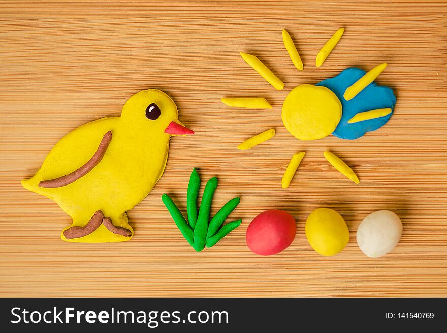 Funny spring Easter image made of plasticine, a yellow chicken, green grass, colorful eggs - red, yellow and white, golden sun shining over blue cloud on wooden background, warm colors