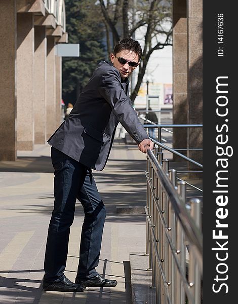 Young businessman leans on metal handrails near the office building with columns. Young businessman leans on metal handrails near the office building with columns