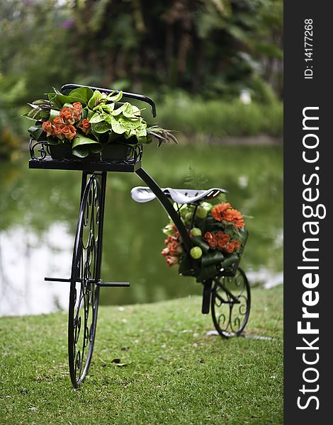 A wrought iron bicycle decorated with flowers on a green grass patch with a pond in the background. A wrought iron bicycle decorated with flowers on a green grass patch with a pond in the background