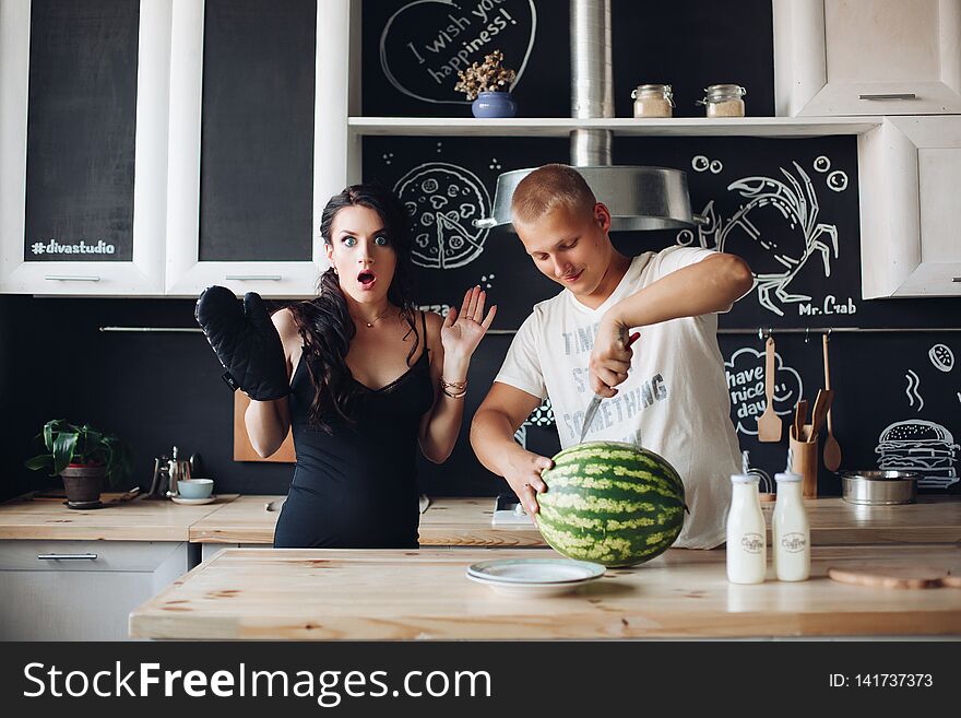 Happy young couple of people standing at kitchen and eating fruits. Beautiful brunette pregnant women holding plate and standing near husband. Handsome men cutting watermelon and sharing it with wife. Happy young couple of people standing at kitchen and eating fruits. Beautiful brunette pregnant women holding plate and standing near husband. Handsome men cutting watermelon and sharing it with wife.