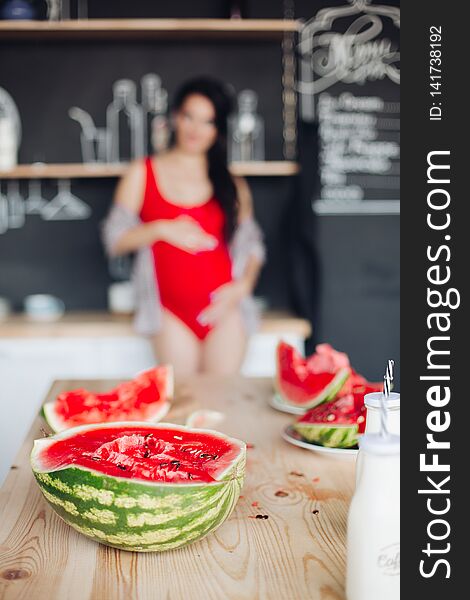 Focus on cut juicy watermelon on wooden table. Unfocused blurred background with unrecognizable pregnant brunette woman in red embracing her belly. Focus on cut juicy watermelon on wooden table. Unfocused blurred background with unrecognizable pregnant brunette woman in red embracing her belly.