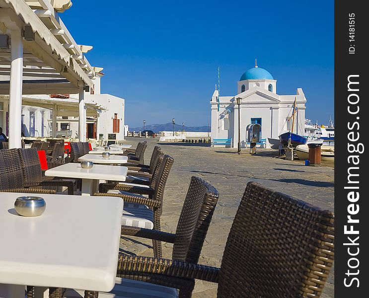 Tables and chairs for the restaurant on the seafront of Mykonos on the background of the church. Tables and chairs for the restaurant on the seafront of Mykonos on the background of the church