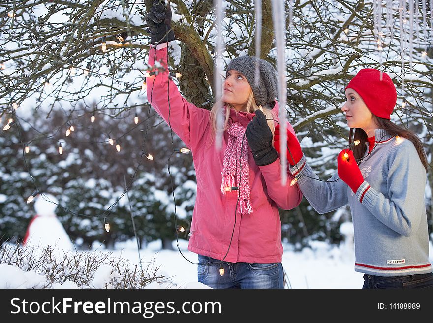 Two Teenage Girls Hanging Fairy Lights In Tree With Icicles In Foreground