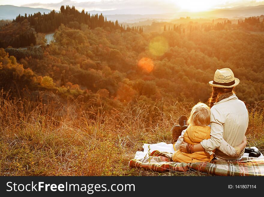 Seen from behind young mother and child hikers in the front of scenery of Tuscany on the sunset looking into the distance while sitting on blanket. Seen from behind young mother and child hikers in the front of scenery of Tuscany on the sunset looking into the distance while sitting on blanket