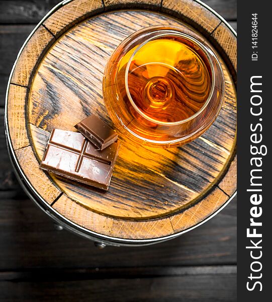 Cognac in a glass with chocolate on a barrel. On black wooden background